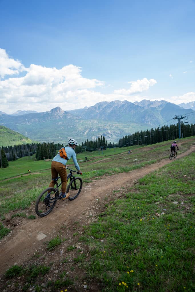mountain bikers ride down a trail on a sunny day with mountains in the background at Purgatory Resort